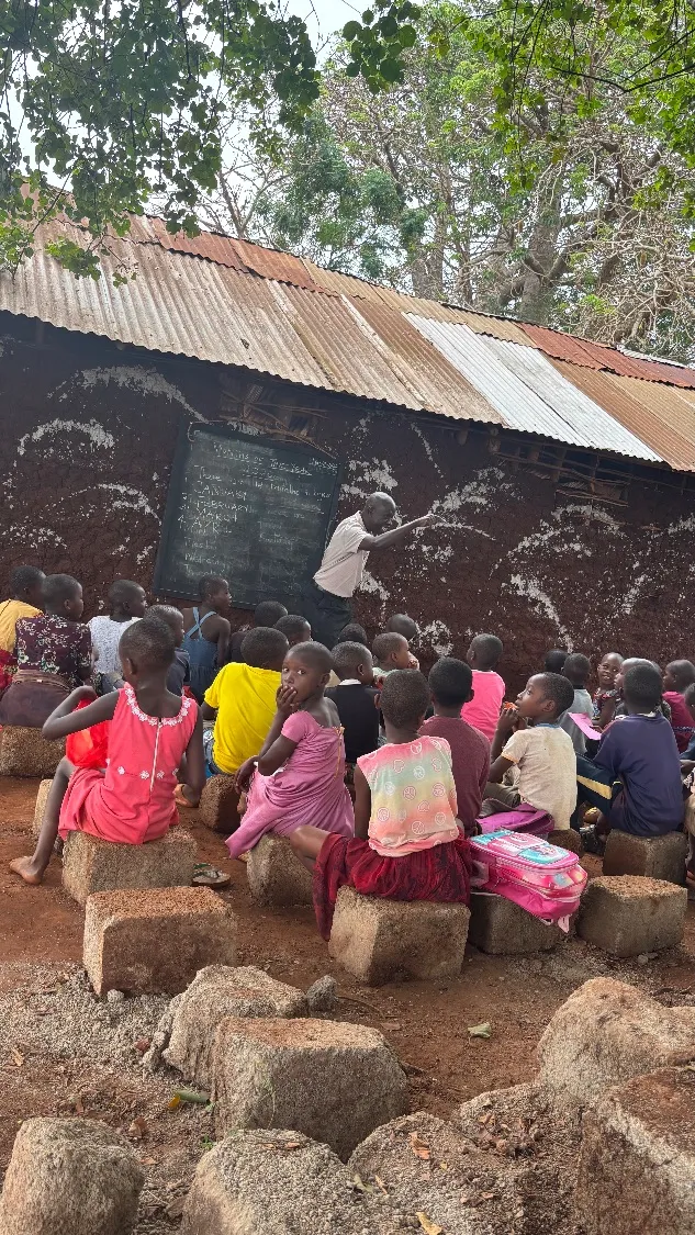 A teacher leads a lesson at an outdoor blackboard mounted on a mud wall, with children seated on stone blocks, in a rural community in Kilifi County, Kenya.