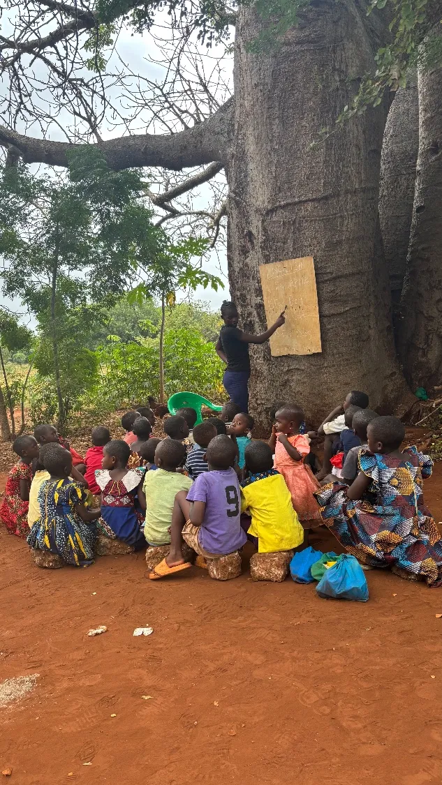 Children sit outdoors beneath a baobab tree as a teacher points to a lesson written on a board, in a rural community in Kilifi County, Kenya.